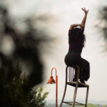 Dancer Bianca Cabrera sits on the edge of a chair, reaching towards the sky.