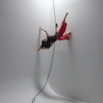 Megan Lowe suspended by rope and harness in the air, dancing in a black top and red pants.