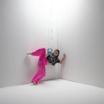 Quinn Dior dancing in a corner with a swinging metal coffee mug, wearing a black top and pink bottoms.