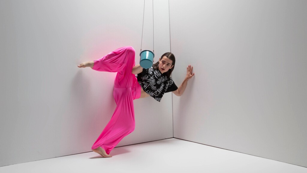 Quinn Dior dancing in a corner with a swinging metal coffee mug, wearing a black top and pink bottoms.
