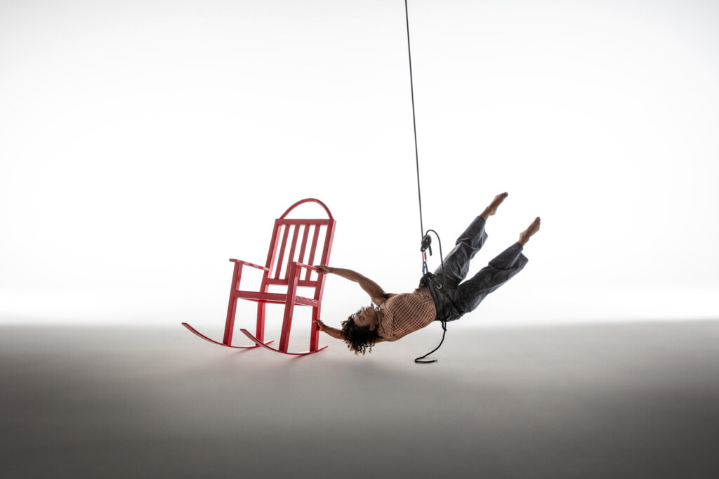 Clarissa Dyas dancing in the air suspended via rope and harness, with a red rocking chair on the ground.